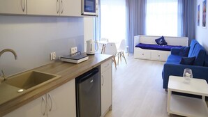 Une cuisine moderne équipée d’un lave-vaisselle, d’un four à micro-ondes et d’une bouilloire sur le plan de travail. On y trouve un coin repas avec une table et des chaises blanches, ainsi qu’un canapé bleu dans le salon.
