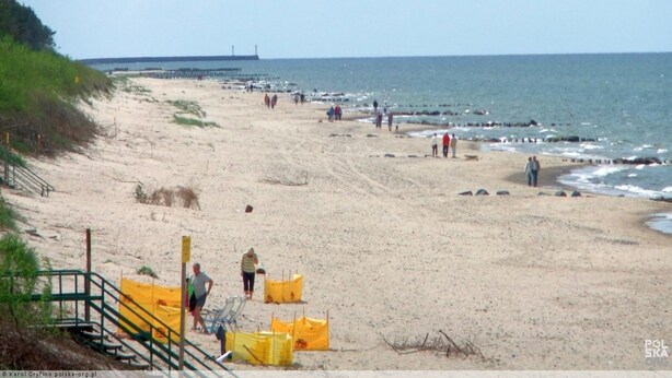 Une plage où l’on voit des gens se promener, une jetée en arrière-plan et des barrières de sécurité jaunes.