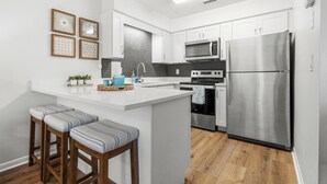 A modern kitchen with white cabinetry, a stainless steel refrigerator, a microwave, and a built-in oven. There are two wooden stools with striped cushions and a small island with a sink and decorative items. - Laguna Beach Community | Steps to Beach • Shared Pool + Pickleball | Gulf Walk (Panama City Beach)