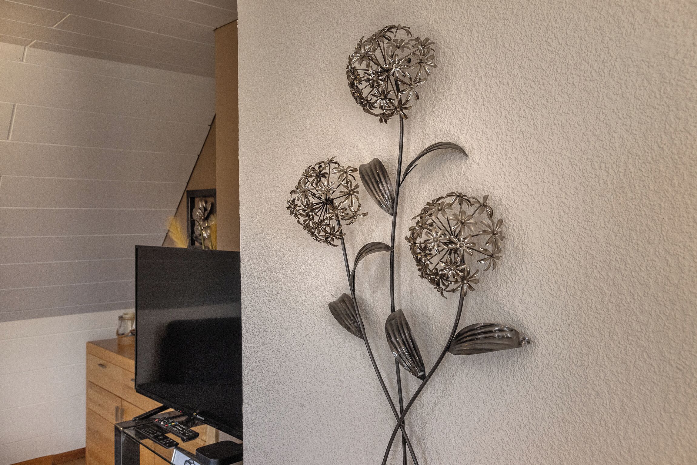 Una sala moderna con televisor de pantalla plana sobre un soporte de madera, una obra de arte metálica decorativa en la pared y una escalera al fondo.