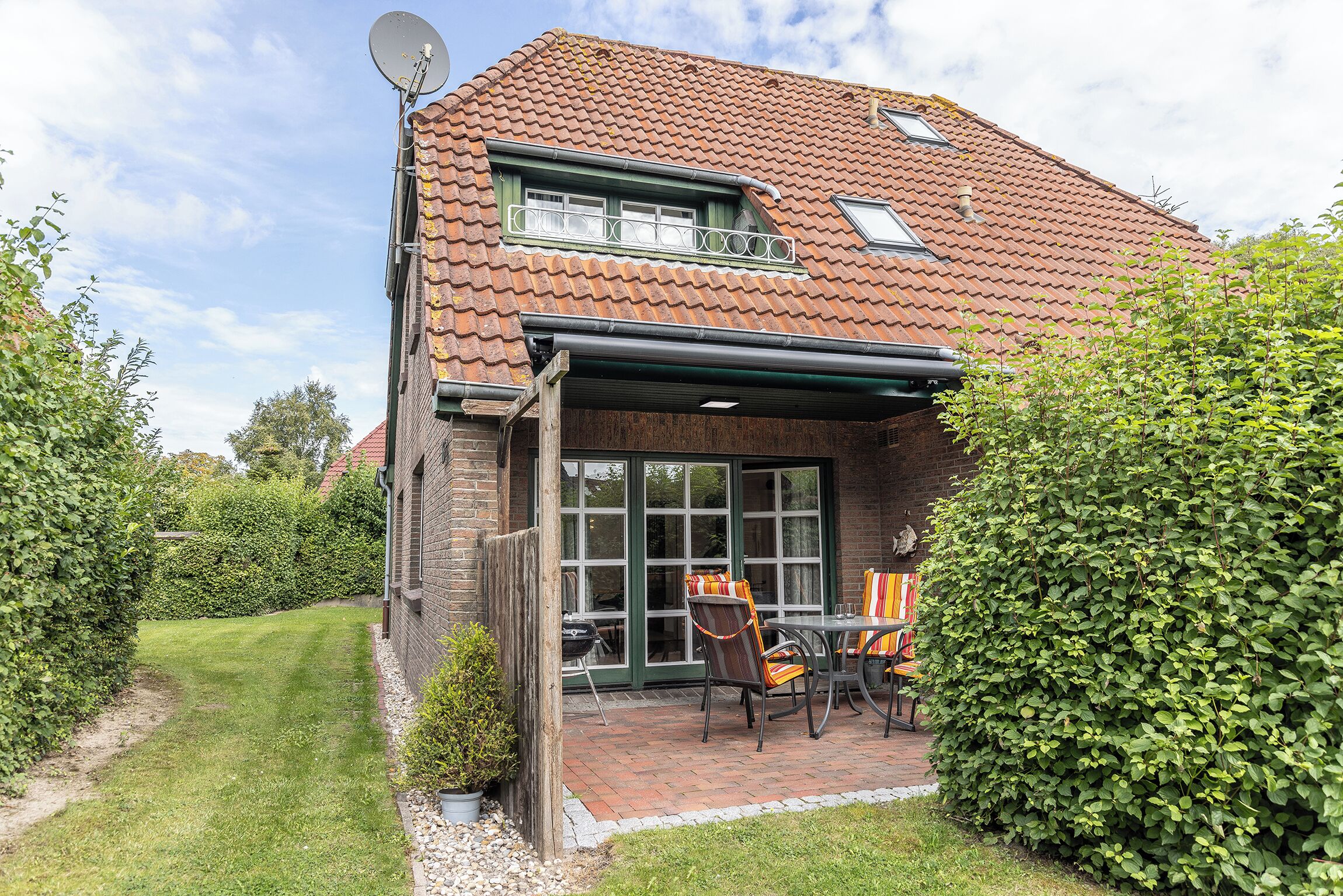 A house with a tiled roof, a satellite dish, a covered patio with chairs, and a well-maintained lawn.