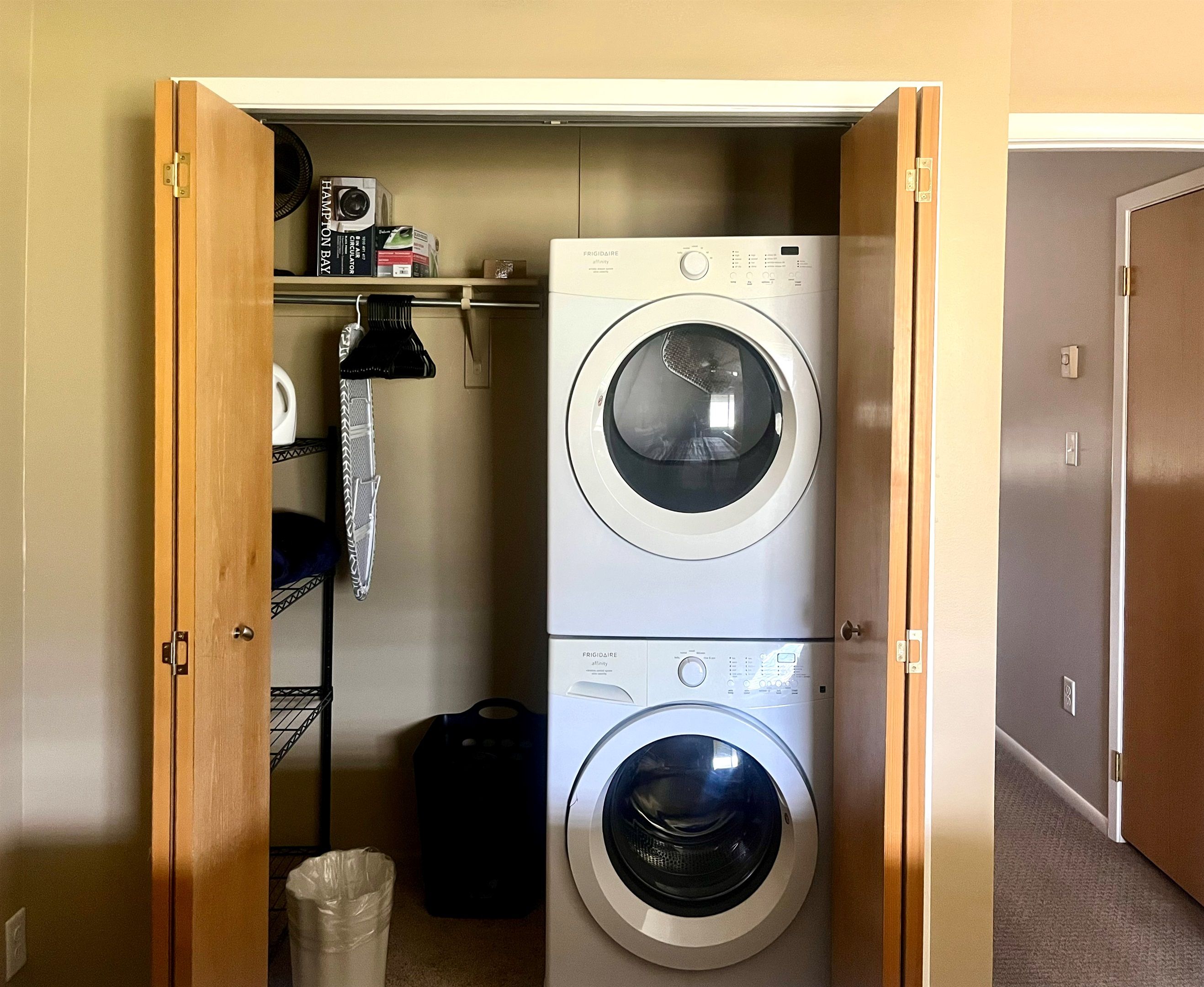 Una sala de lavandería con dos lavadoras de carga frontal apiladas, un estante con libros y una cámara, y un cubo de basura.