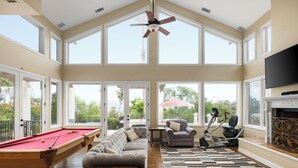 Amplia sala de estar con mesa de billar, asientos cómodos, equipo de ejercicio y grandes ventanales con vistas al exterior.