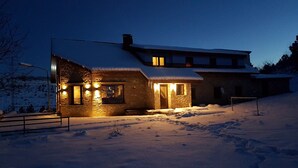 A stone building with a snow-covered roof, illuminated windows, and a snow-covered ground. - katafigio seli (Veroia)