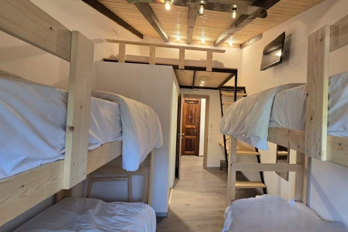 Quarto com beliches, vigas de madeira e televisão fixada na parede.