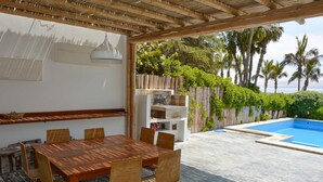 An outdoor dining area with a wooden table and chairs, a built-in grill, and a view of a swimming pool and palm trees. - w* | Modern 5BR with Private Pool in Paracas (Paracas)