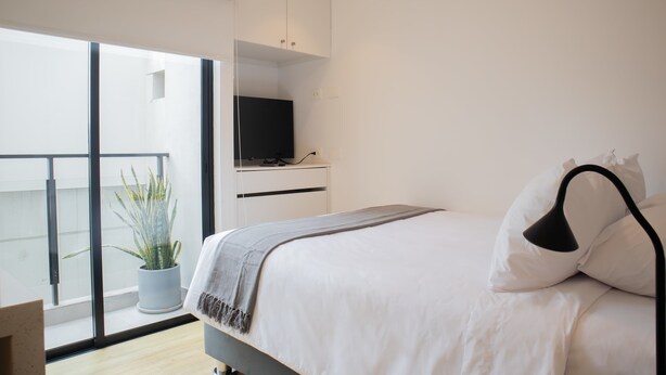 A modern bedroom with a bed, a TV on a stand, a potted plant, and a balcony with a view. - w* | Seaside Studio near Malecon in Miraflores (Miraflores)