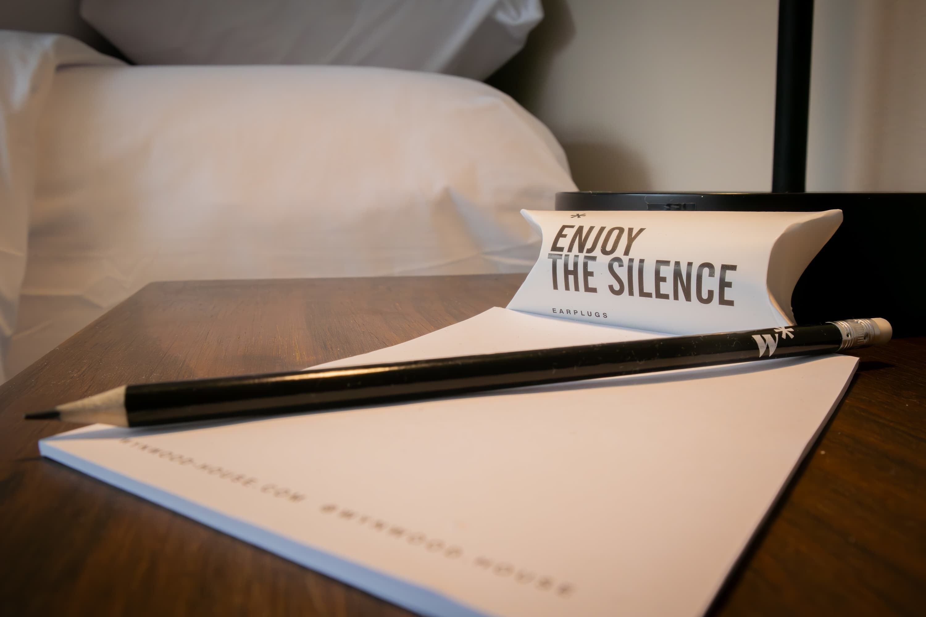 A hotel room with a bed, a bedside table with a notepad, a pen, and a sign that reads "ENJOY THE SILENCE EARPLUGS".