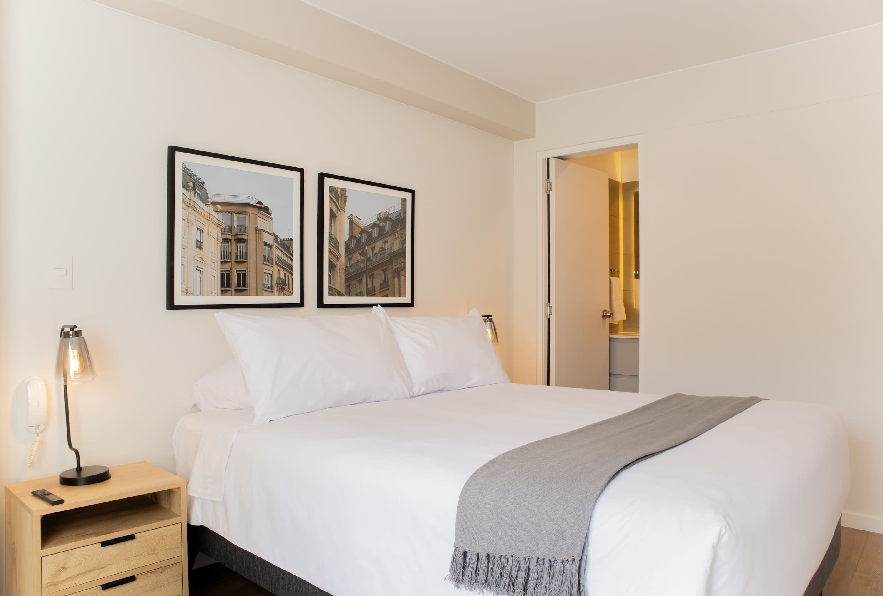 Un lit bien fait, avec du linge de lit blanc et un plaid gris, une table de chevet avec une lampe, et deux tableaux encadrés accrochés au mur.