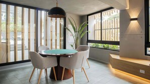 Un comedor moderno con una mesa redonda, cuatro sillas, una lámpara colgante y un ventanal grande con persianas verticales.