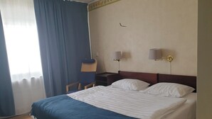 Quarto de hotel com cama, uma cadeira, duas luminárias de cabeceira e janela com cortinas.