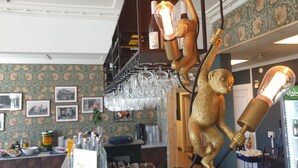 Uma área de bar com balcão, copos pendurados no teto e um macaco dourado decorativo segurando uma lâmpada.