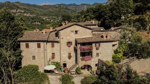 A stone building with multiple windows and balconies, surrounded by greenery and hills.