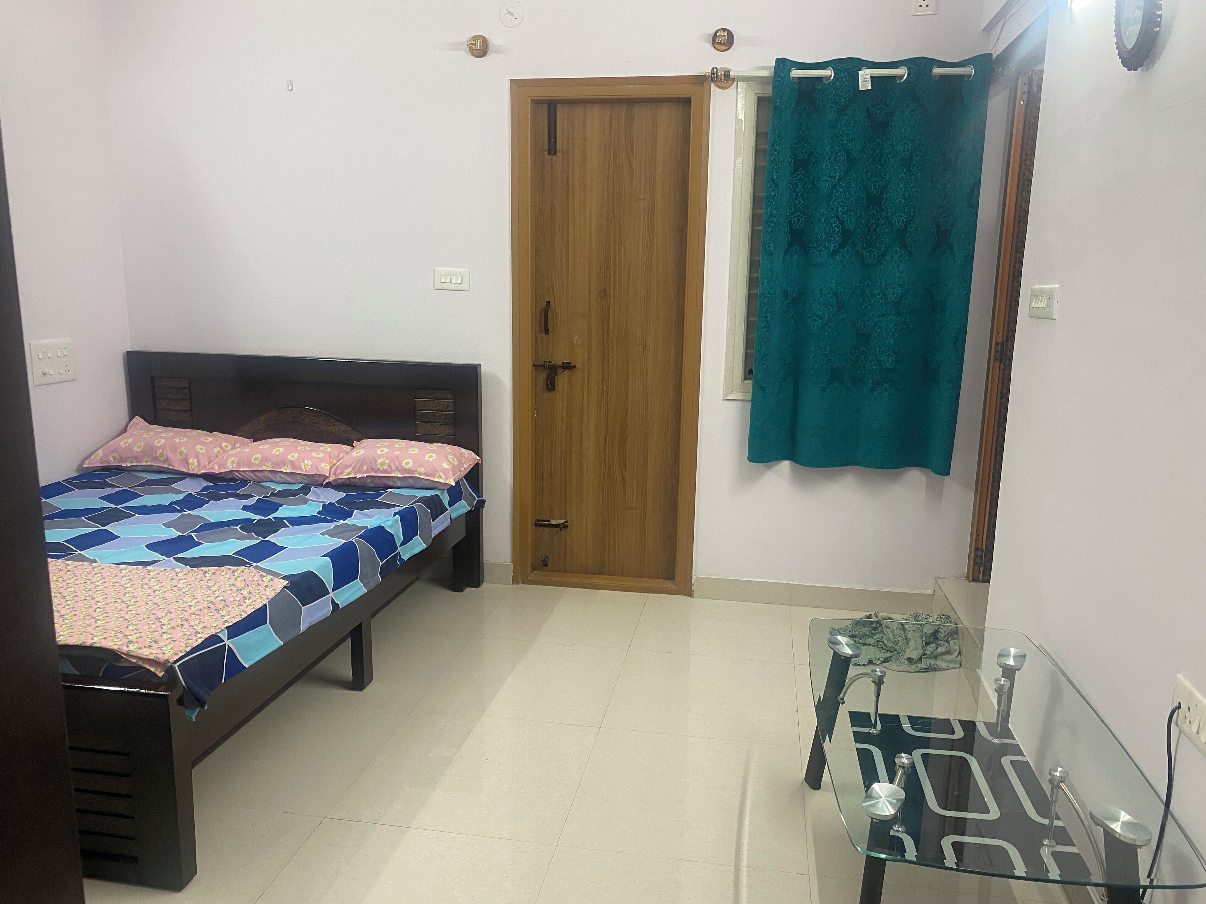 Un dormitorio con una cama, una puerta de madera, una ventana con cortina y una pequeña mesa con superficie de vidrio.