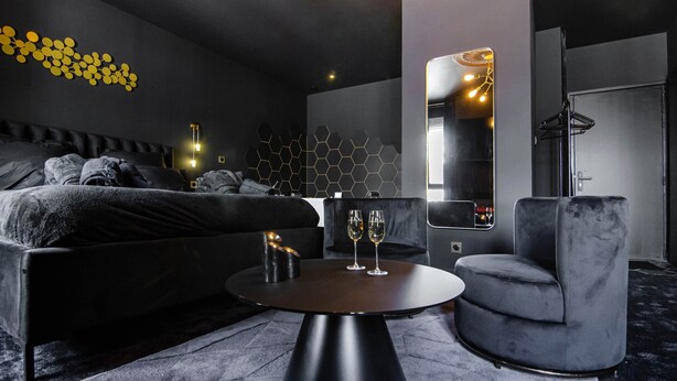 A modern hotel room with a bed, a round table with two glasses, a grey armchair, and a wall with a geometric light fixture.