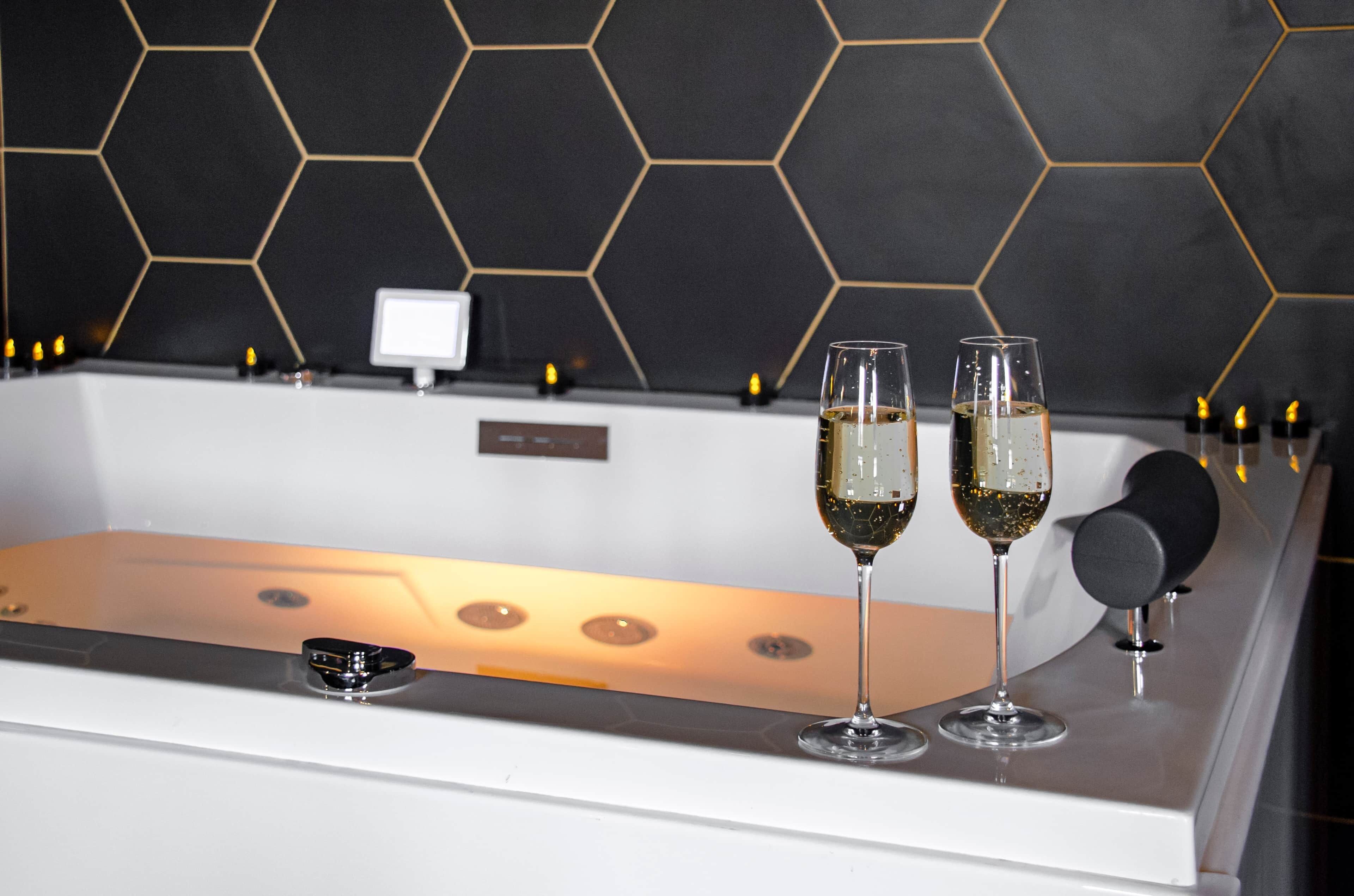 Ein modernes Badezimmer mit einer Wand aus schwarzen, sechseckigen Fliesen, einer weißen Badewanne und zwei mit einer klaren Flüssigkeit gefüllten Sektgläsern.