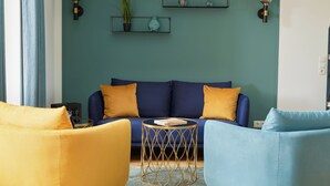 A living room with a blue sofa, yellow and blue armchairs, a round coffee table, and wall-mounted shelves with decorative items. - Completion 2025: Exclusive vacation apartment in prime location near the beach! (Sankt Peter-Ording)