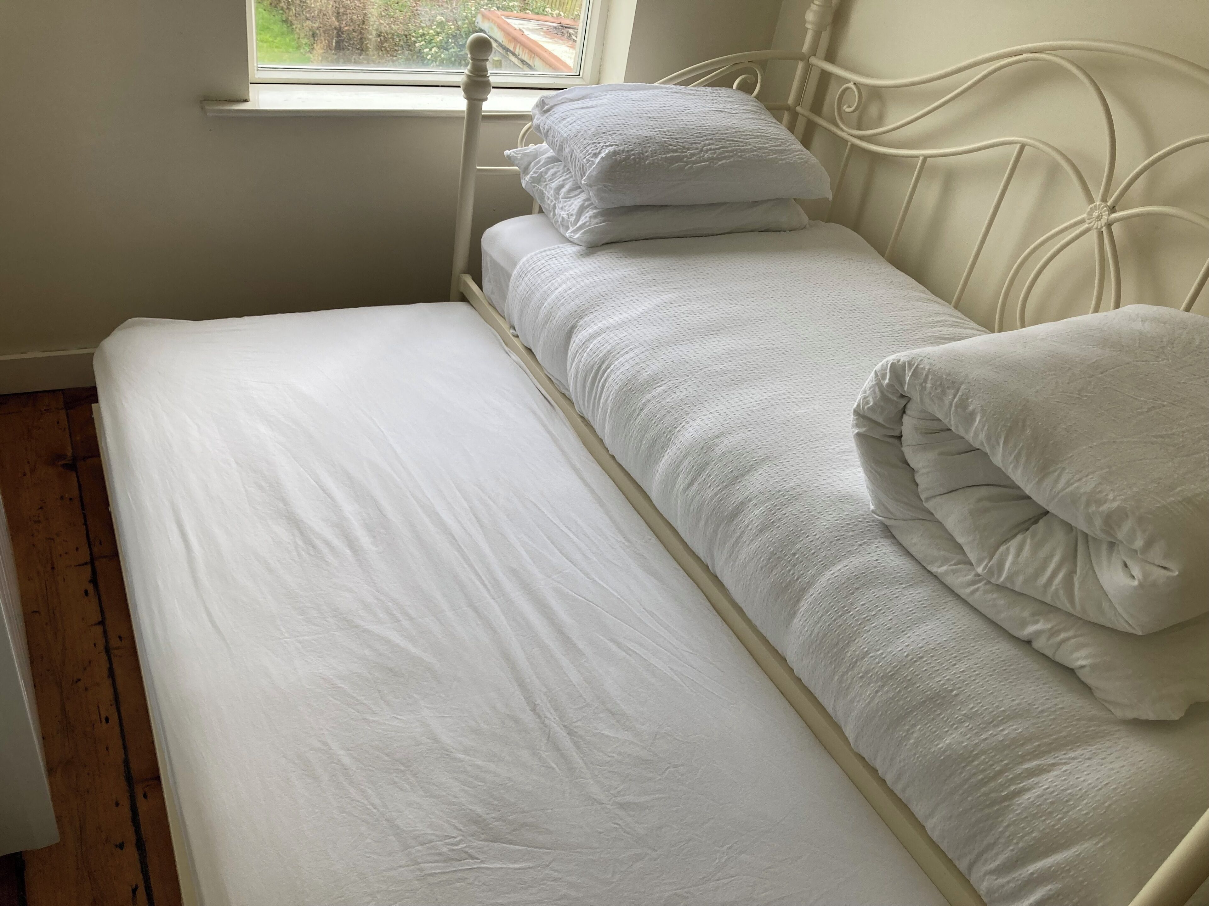 A single bed with white bedding and pillows, a window with a view of greenery, and a wooden floor.