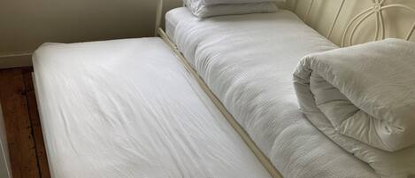 A single bed with white bedding and pillows, a window with a view of greenery, and a wooden floor.