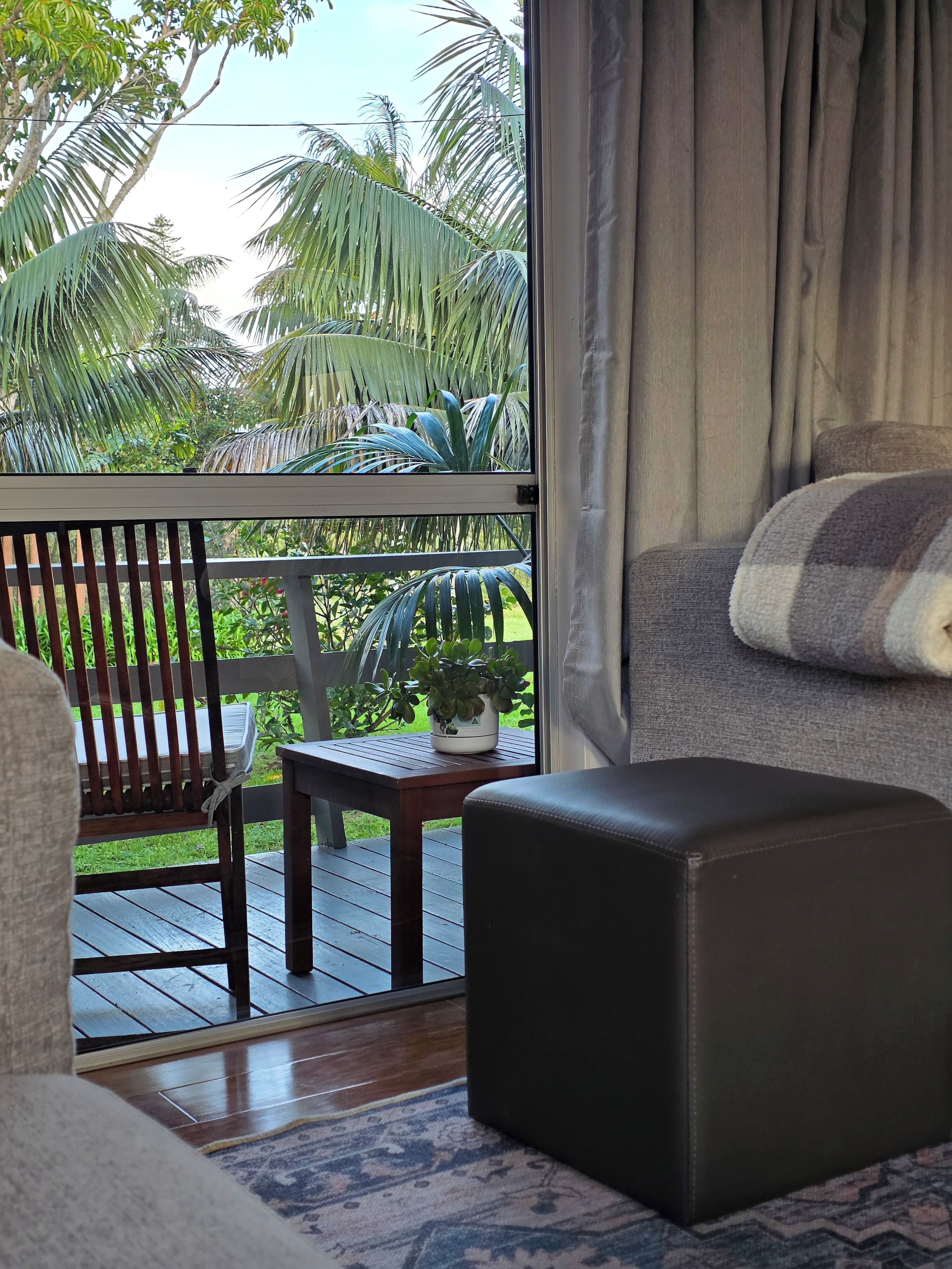 Une pièce avec vue sur une terrasse en bois et des palmiers, un canapé gris avec un coussin à rayures et une petite table sur laquelle est posée une plante.