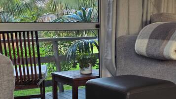Une pièce avec vue sur une terrasse en bois et des palmiers, un canapé gris avec un coussin à rayures et une petite table sur laquelle est posée une plante.