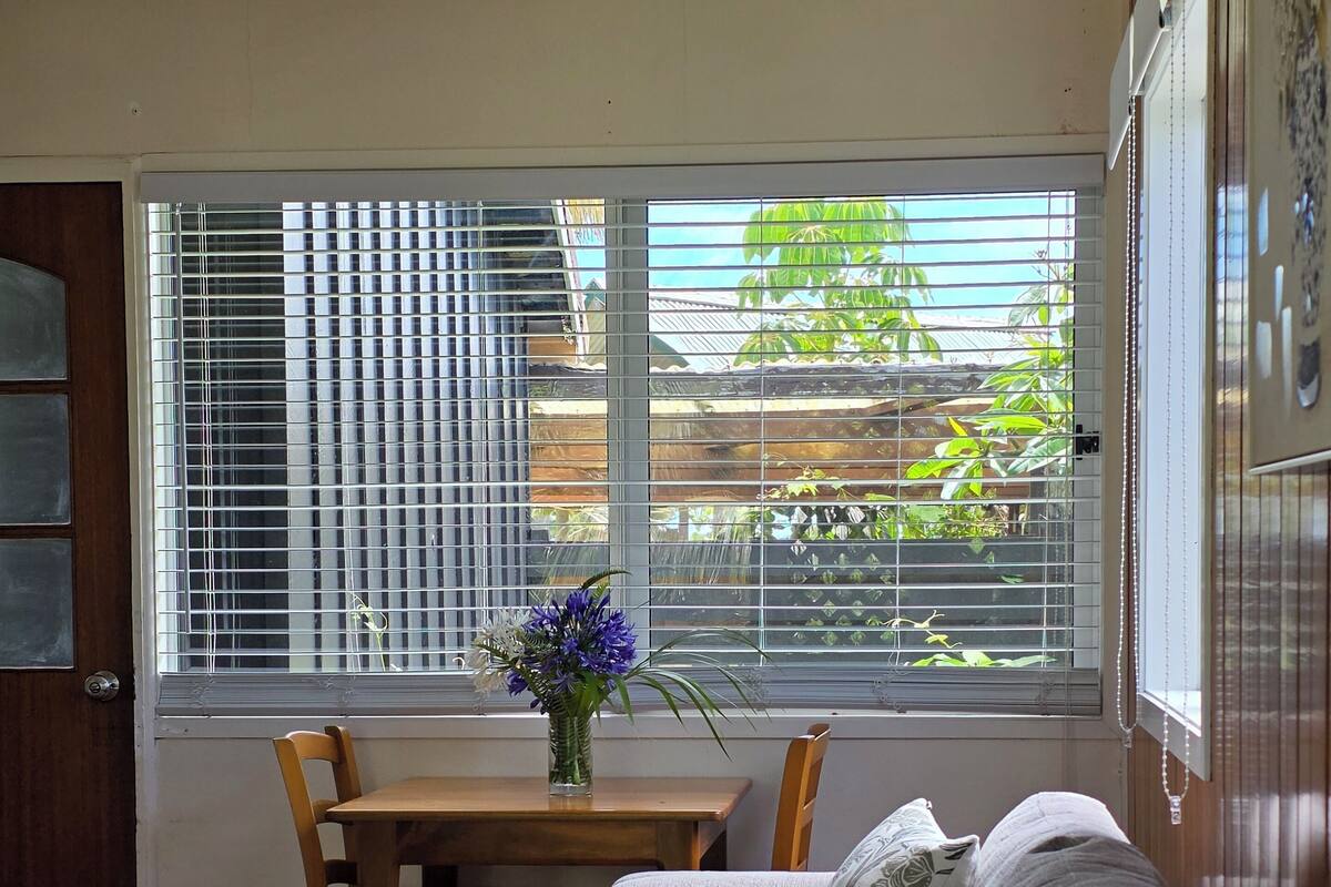A living room with a sofa, a table with a vase of flowers, a dining table with chairs, and a view through the window.