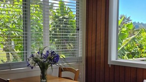 A wooden dining table with a vase of flowers, a chair, and a window with blinds overlooking a garden. - Fletcher Christian Apartments - Isabella Suites (Burnt Pine)