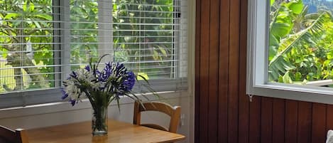 Una mesa de comedor de madera con un florero, una silla y una ventana con persianas que da a un jardín.
