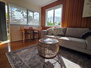 A living room with a sofa, a coffee table, a dining table with chairs, and a view of the outdoors through the windows. - Fletcher Christian Apartments - Isabella Suites (Burnt Pine)