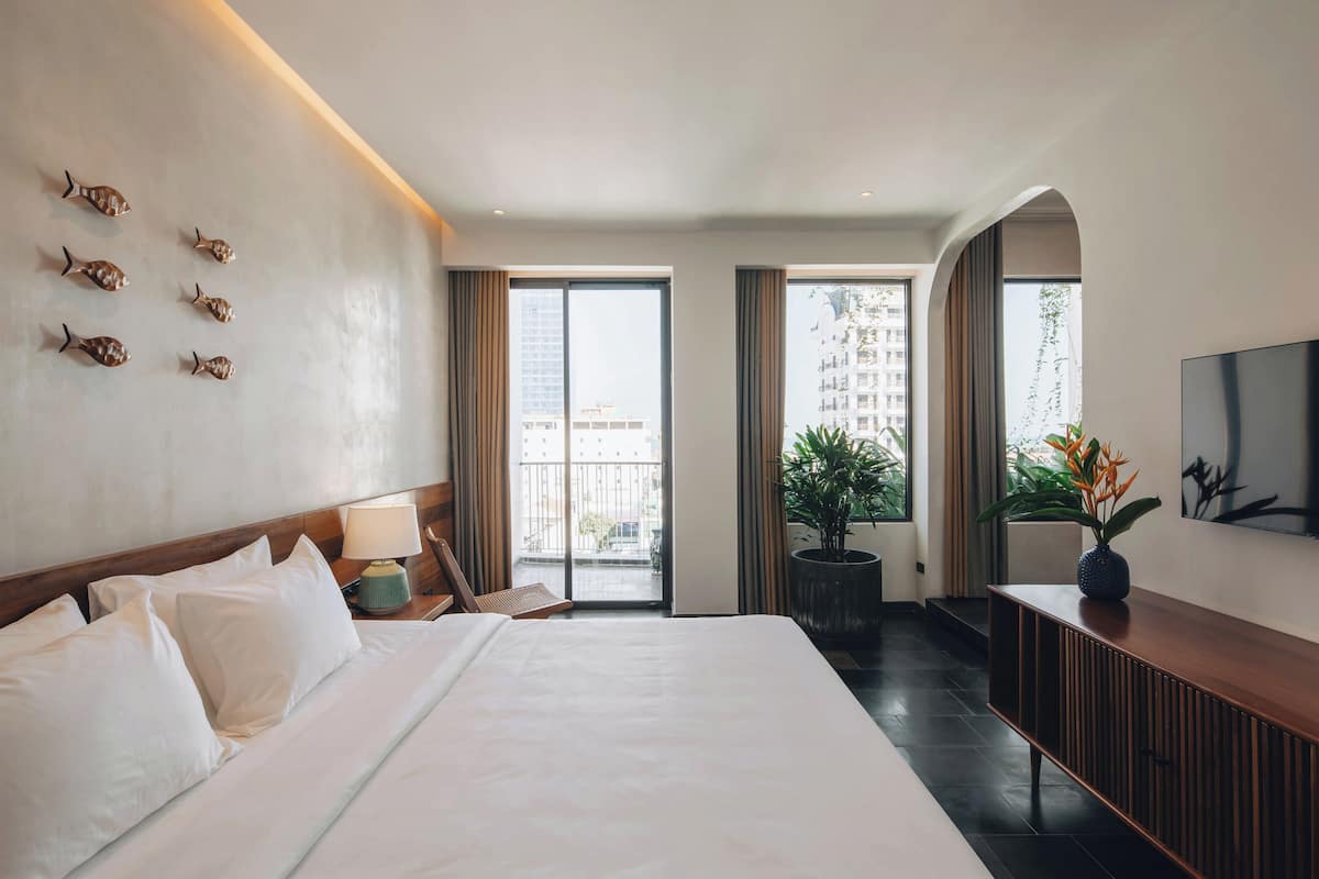 A hotel room with a large bed, a nightstand with a lamp, a TV on the wall, and a balcony with a view of buildings.