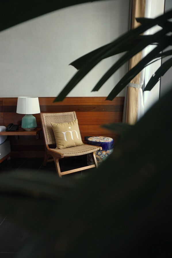 A room with a wooden headboard, a wicker chair with a pillow, a small table with a lamp, and a decorative bowl.