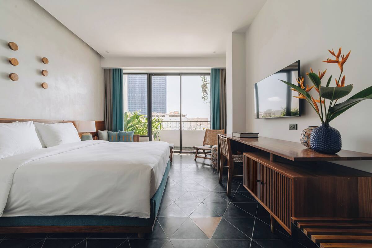 A modern hotel room with a large bed, a desk, and a balcony with a view of the city.