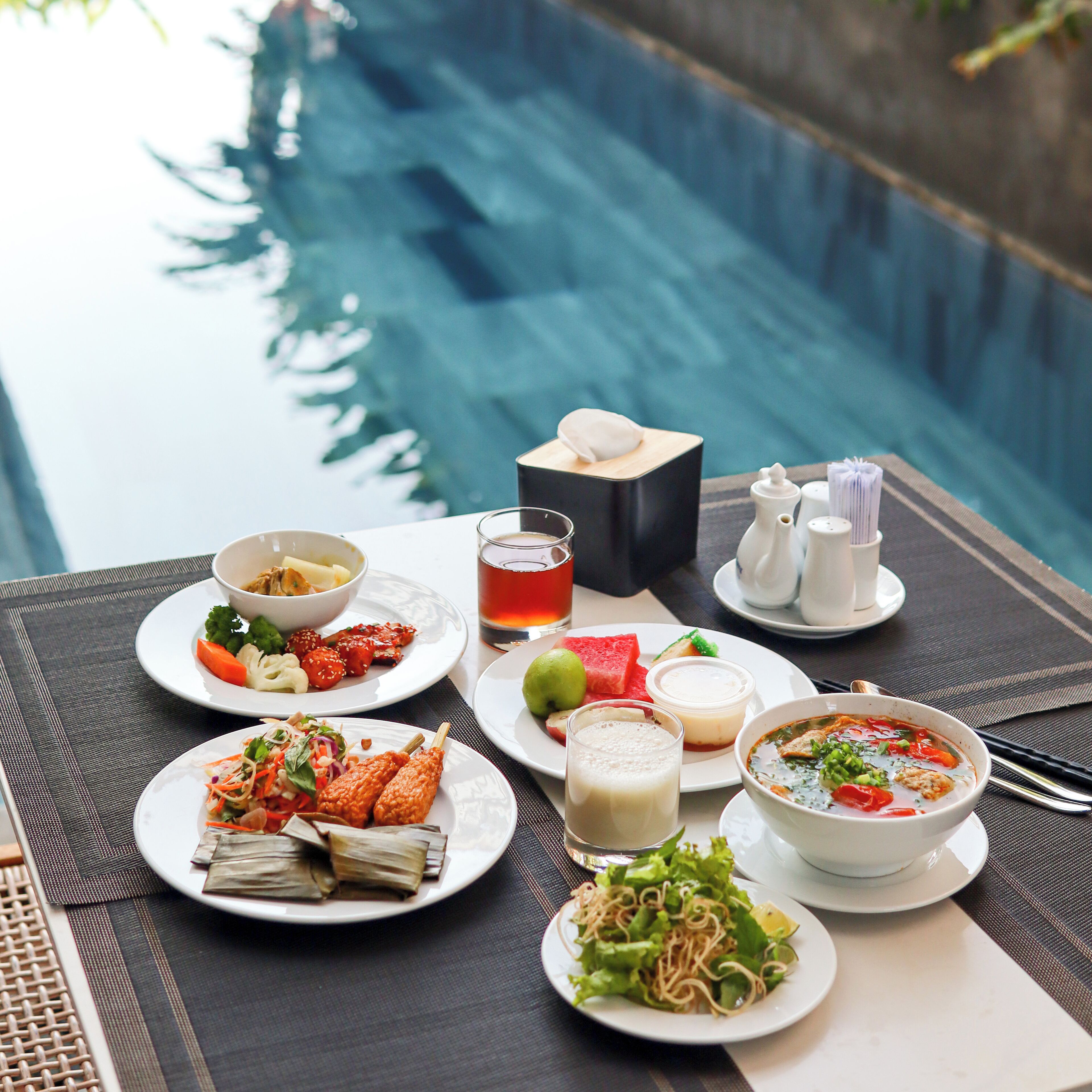 A dining table with various dishes, a beverage, and condiments, set against a poolside view.