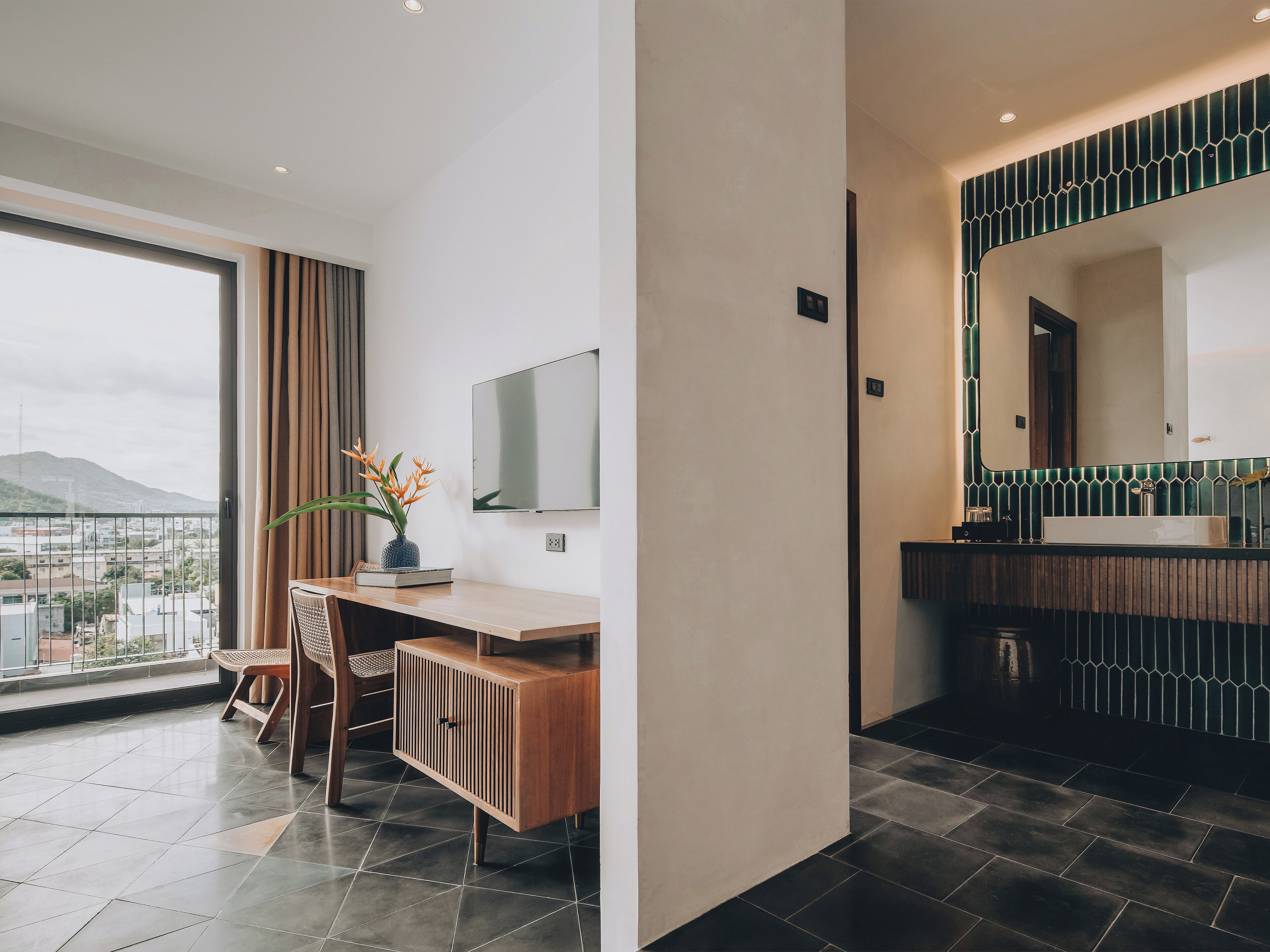 Quarto de hotel moderno com uma janela grande, uma escrivaninha com cadeira, televisão de tela plana e banheiro com espelho amplo e azulejos escuros.
