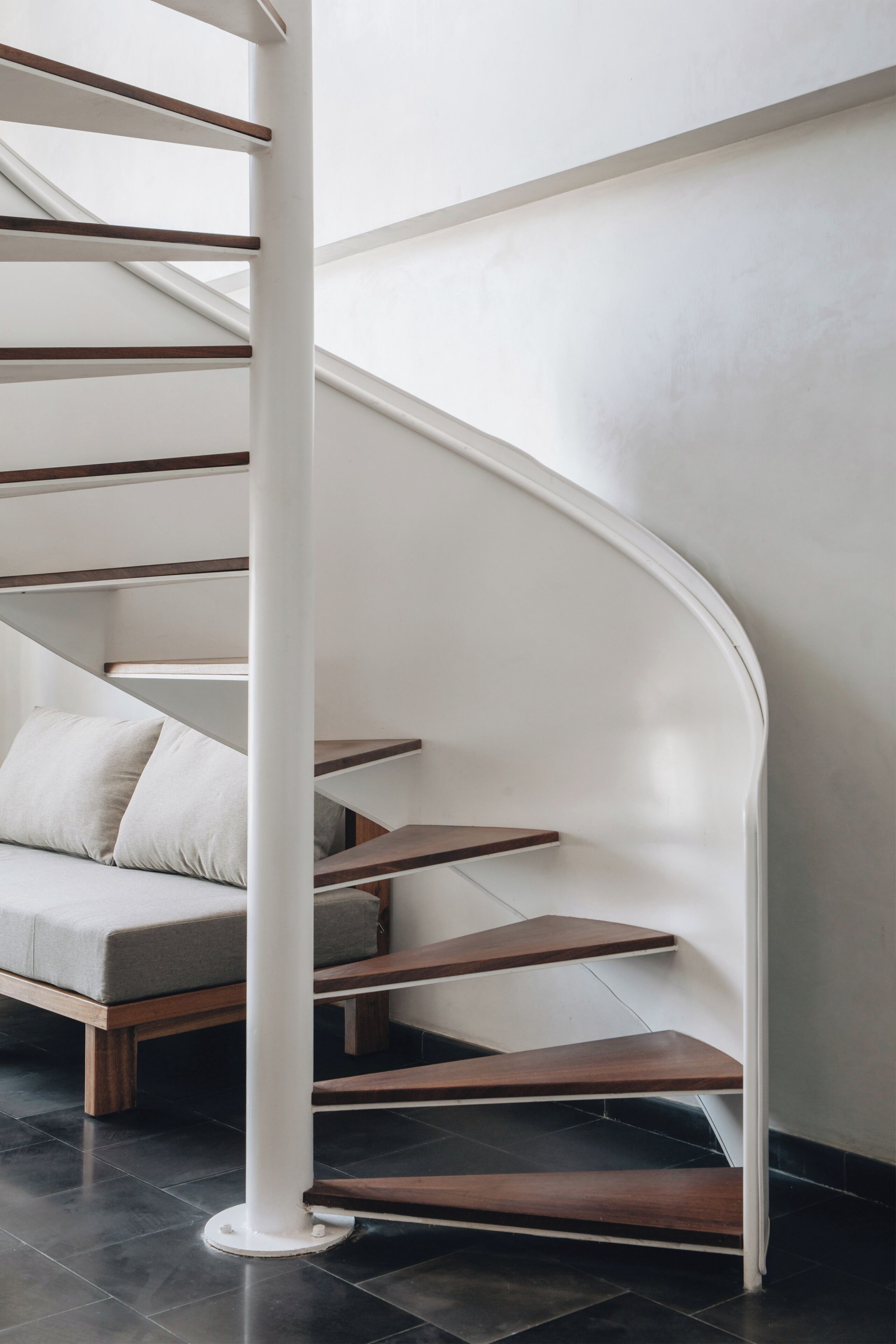 A spiral staircase with a white column, wooden steps, and a grey sofa with cushions.