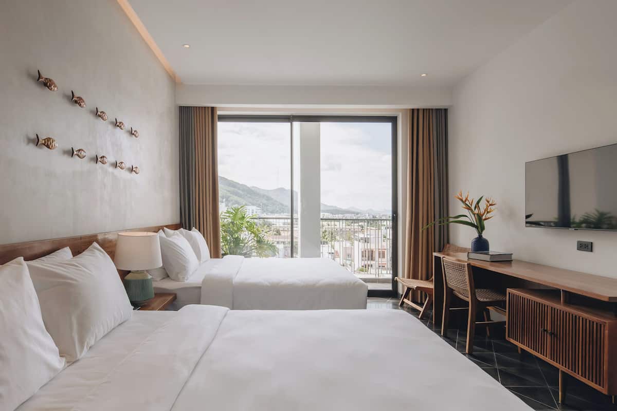 A hotel room with two beds, a desk, a TV, and a view of the city and mountains.