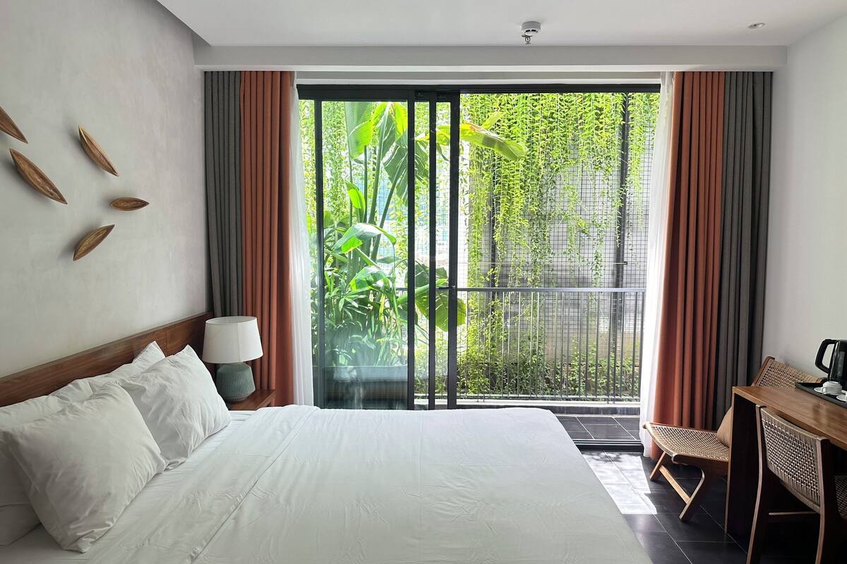 A hotel room with a bed, a desk, a chair, a lamp, and a view of greenery through a sliding door.