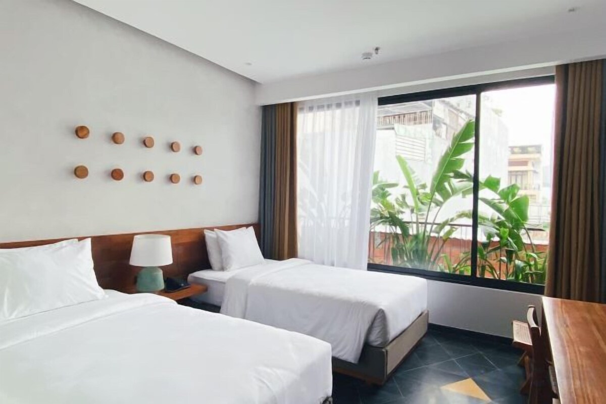 Habitación de hotel con dos camas, una ventana con cortinas, una planta afuera y un escritorio de madera.