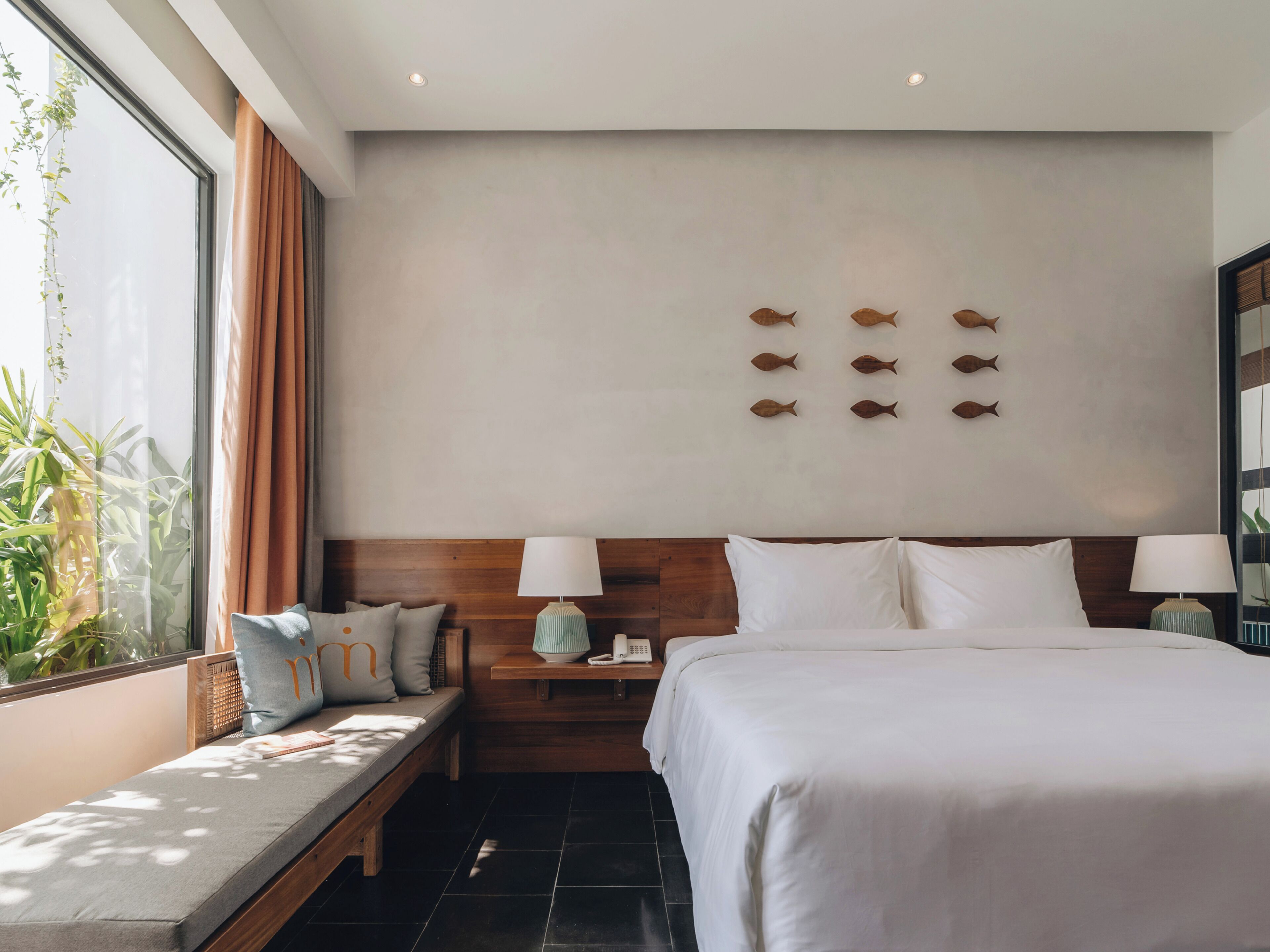 A hotel room with a large bed, bedside tables with lamps, a wooden headboard, a bench with cushions, and a wall with fish decorations.