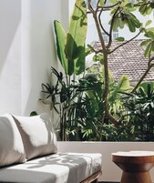 A modern living room with a sofa and a wooden side table, a large window with a view of a tiled roof and lush greenery outside.