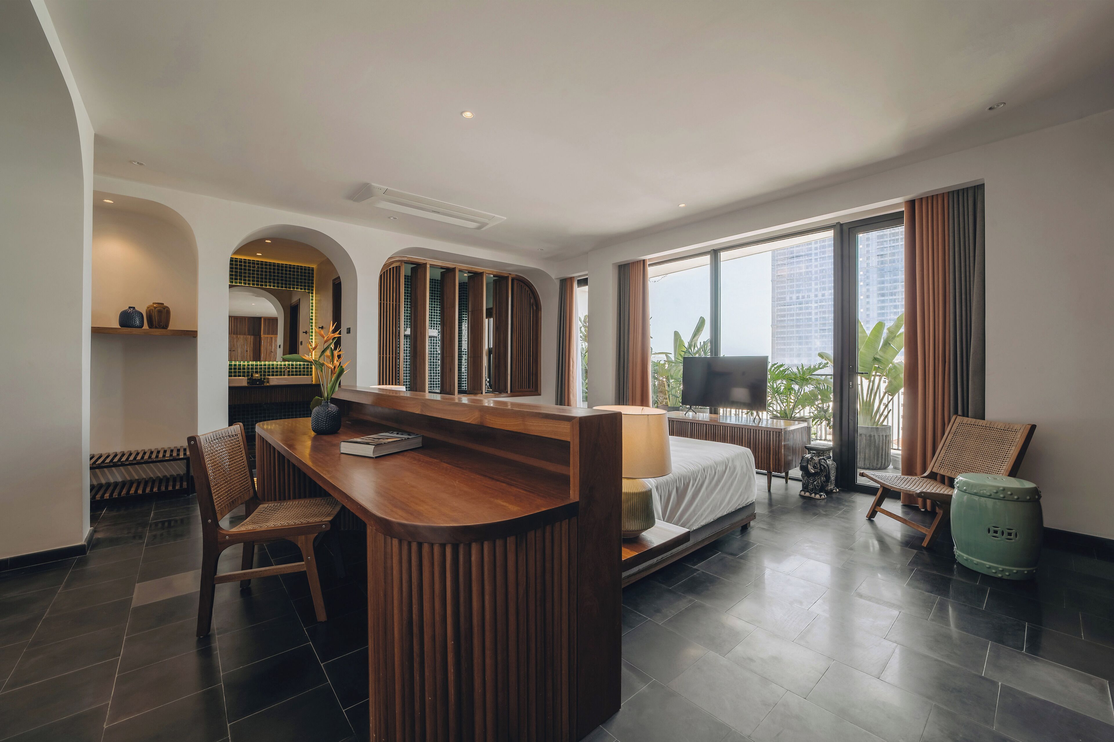 A modern hotel room with a wooden desk, a bed, a TV, and a view of the city.