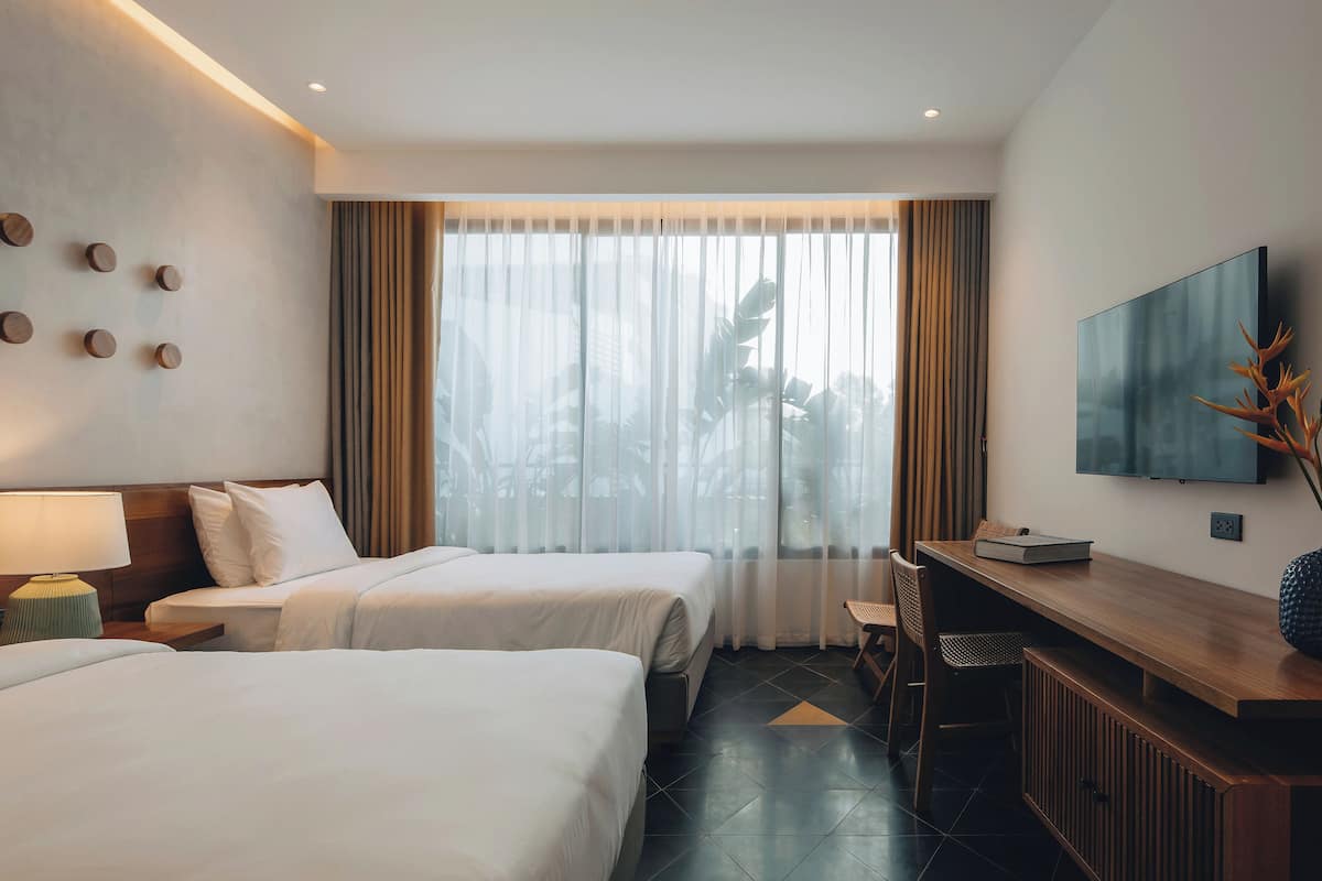 A hotel room with two beds, a desk, a TV, and a large window with curtains.