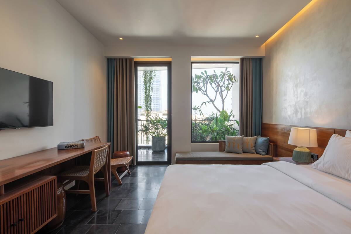 A hotel room with a bed, a desk, a TV, a window with a view of greenery and buildings, and a balcony.