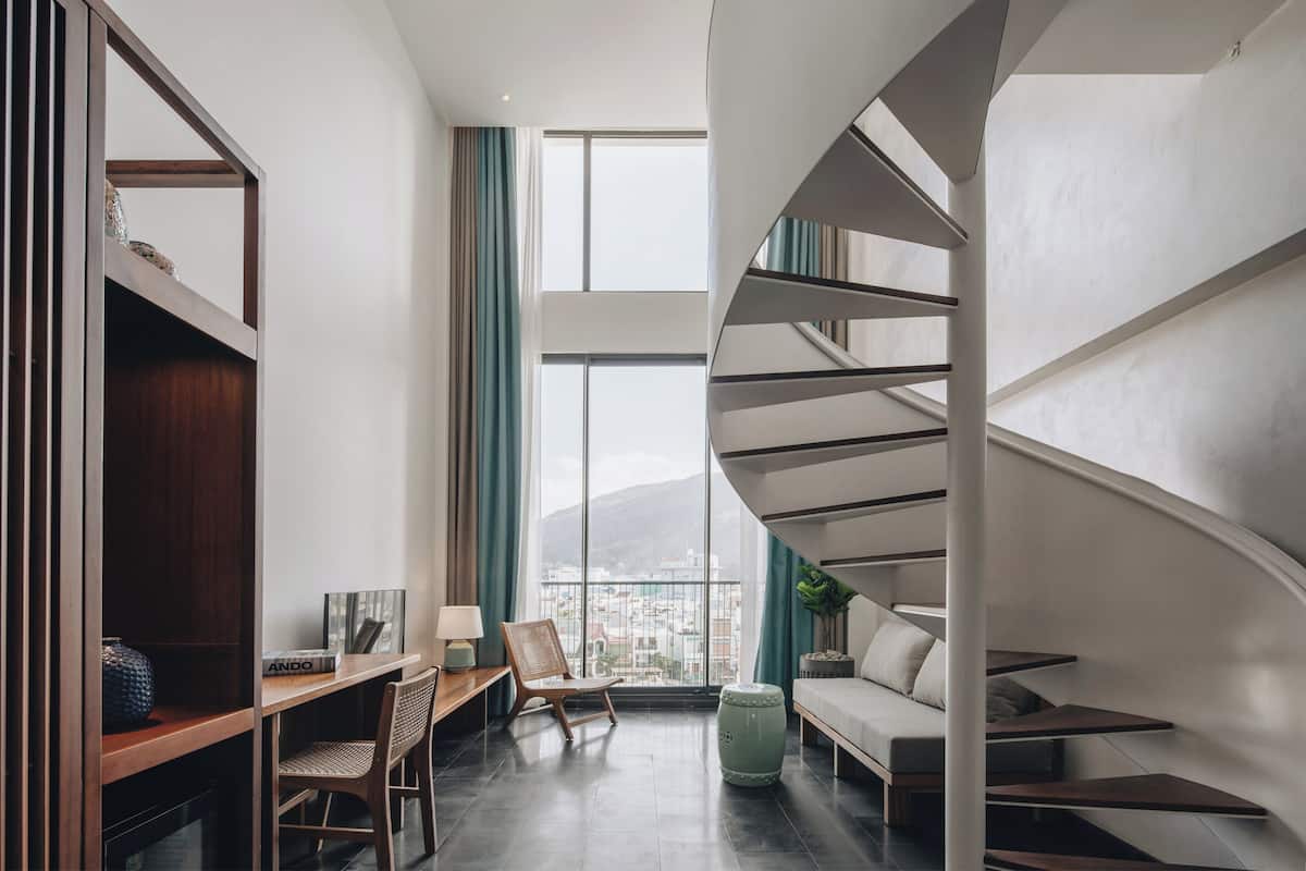 A modern interior with a spiral staircase, a desk with a chair, a sofa, and a large window with a city view.