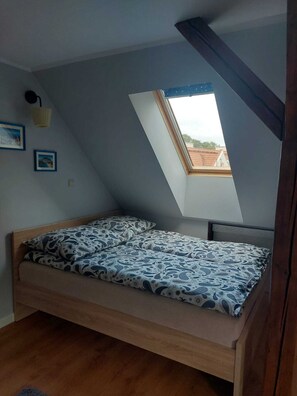 Ein Zimmer mit schrägen Wänden, einem Bett mit gemusterten Betttextilien, einem kleinen Fenster und einem gerahmten Bild an der Wand.