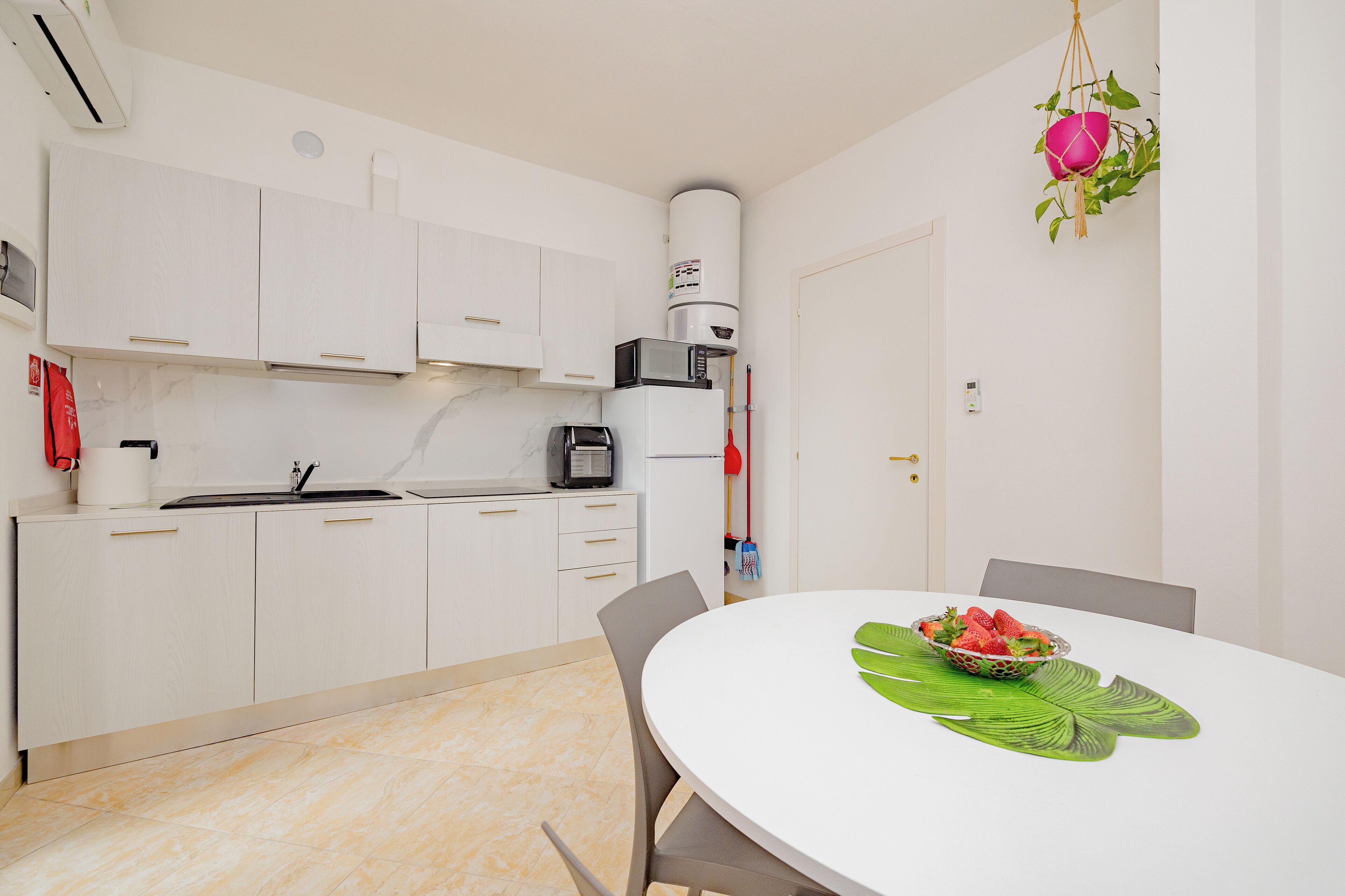 A modern kitchen with white cabinets, a microwave, and a refrigerator. There is a dining table with a bowl of strawberries and a green leaf placemat.