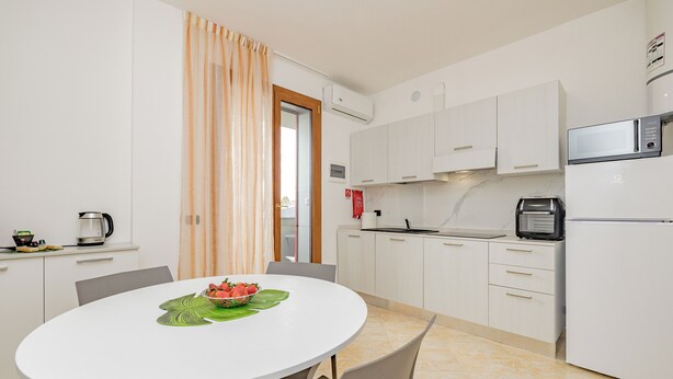 A modern kitchen with white cabinets, a microwave, and a refrigerator. There is a dining table with chairs and a bowl of fruit. - Girasole (A5) | Three-room apartment with two bathrooms | Rho Fiera | Malpensa | Milan (Cesate)