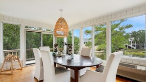 A dining area with a round table, white upholstered chairs, a wicker pendant light, and large windows offering a view of trees and a body of water. - Sonny Pointe On the Creek, Steps away from the Beach, Pet Friendly (Georgetown)