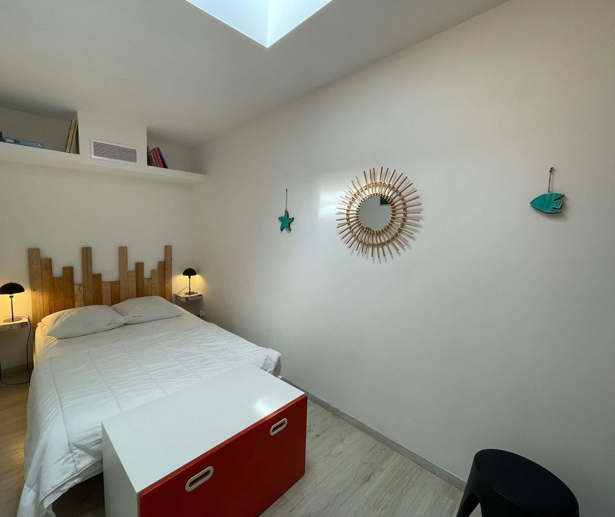 A bedroom with a bed, bedside lamps, a red storage unit, a wall-mounted mirror, and a star decoration.
