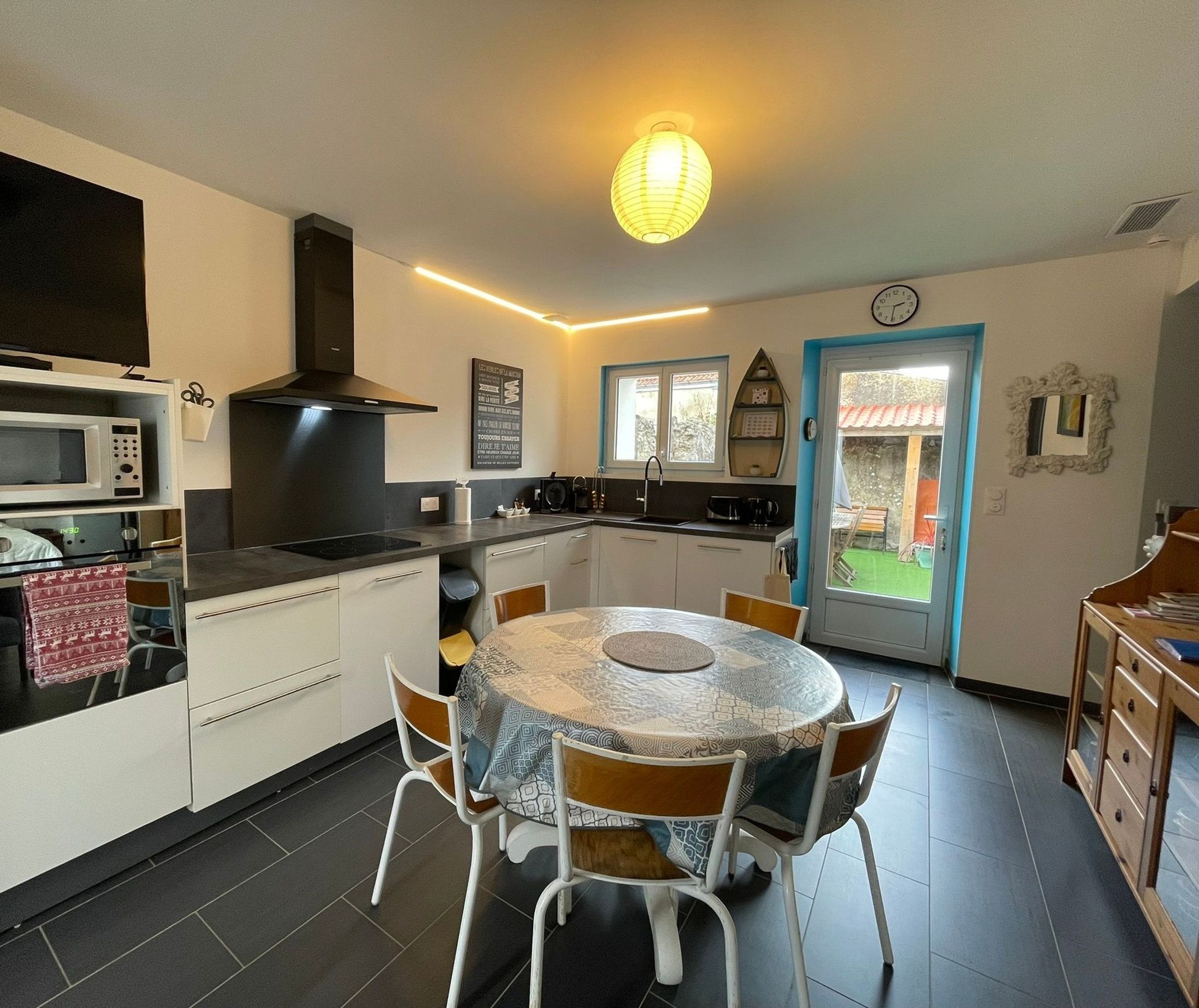 A compact kitchen with a microwave, stove, and sink. There is a dining table with chairs and a clock on the wall.